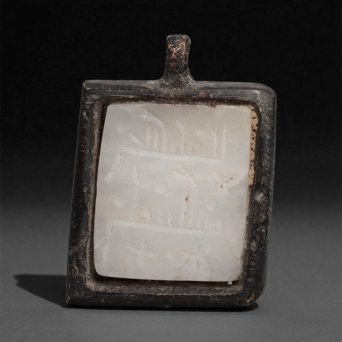 An Islamic Bronze Amulet with Carved Agate Insert, 10th-13th Century CE, 47LP: Formed as a rectangular bronze frame with an integral suspension loop, the amulet houses a fitted plaque of white agate carved in shallow relief. The stone bears linear and dotted motifs arranged in h