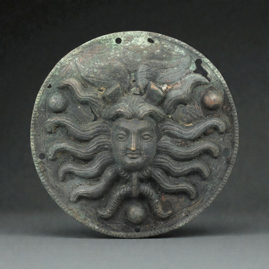 A Greco-Roman Bronze Roundel with Gorgoneion, 1st-3rd Century CE, 78LP: Cast in bronze as a circular applique, the roundel is worked in high relief with a gorgoneion featuring a frontal human face framed by radiating serpentine locks. The face is modeled with gently arche