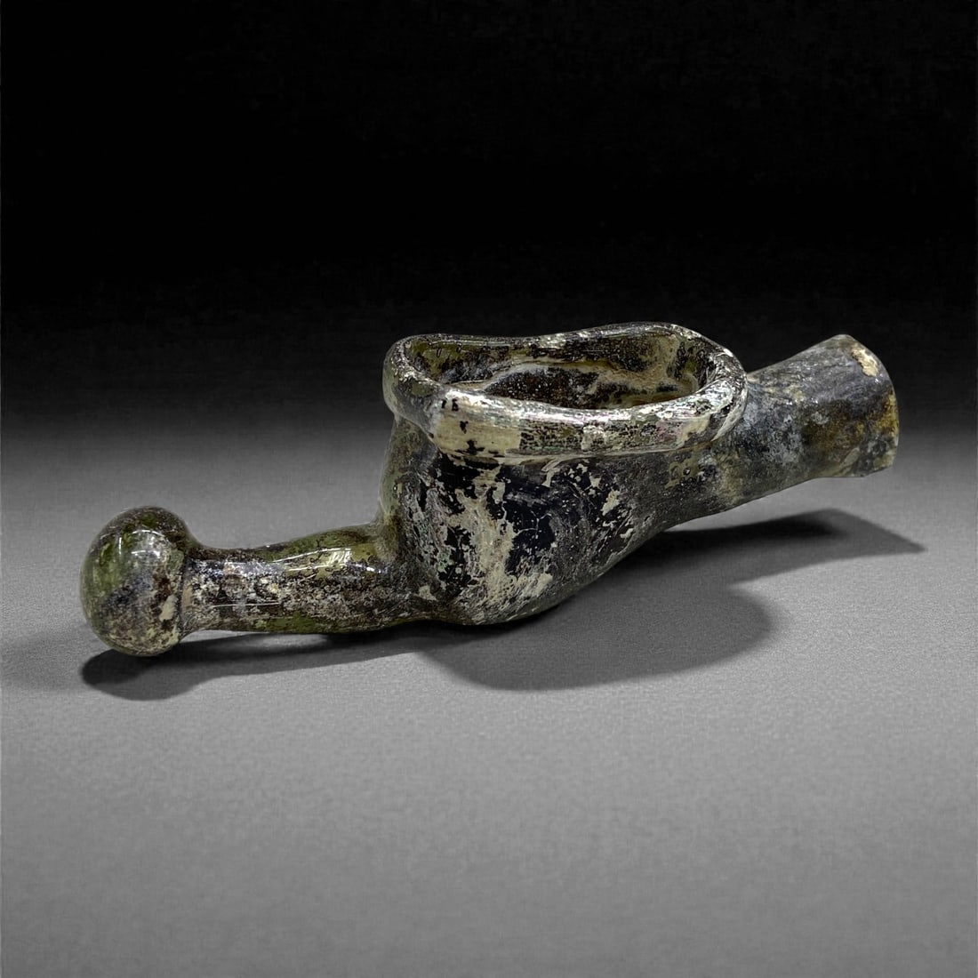 A Roman Levantine Glass Spouted Lamp, 1st-2nd Century CE, 36 g, 28AA: Blown from pale green glass now darkened by heavy weathering, the object is formed with an elongated tubular body that widens into a shallow, oval bowl and terminates in a rounded nozzle. The rim of t