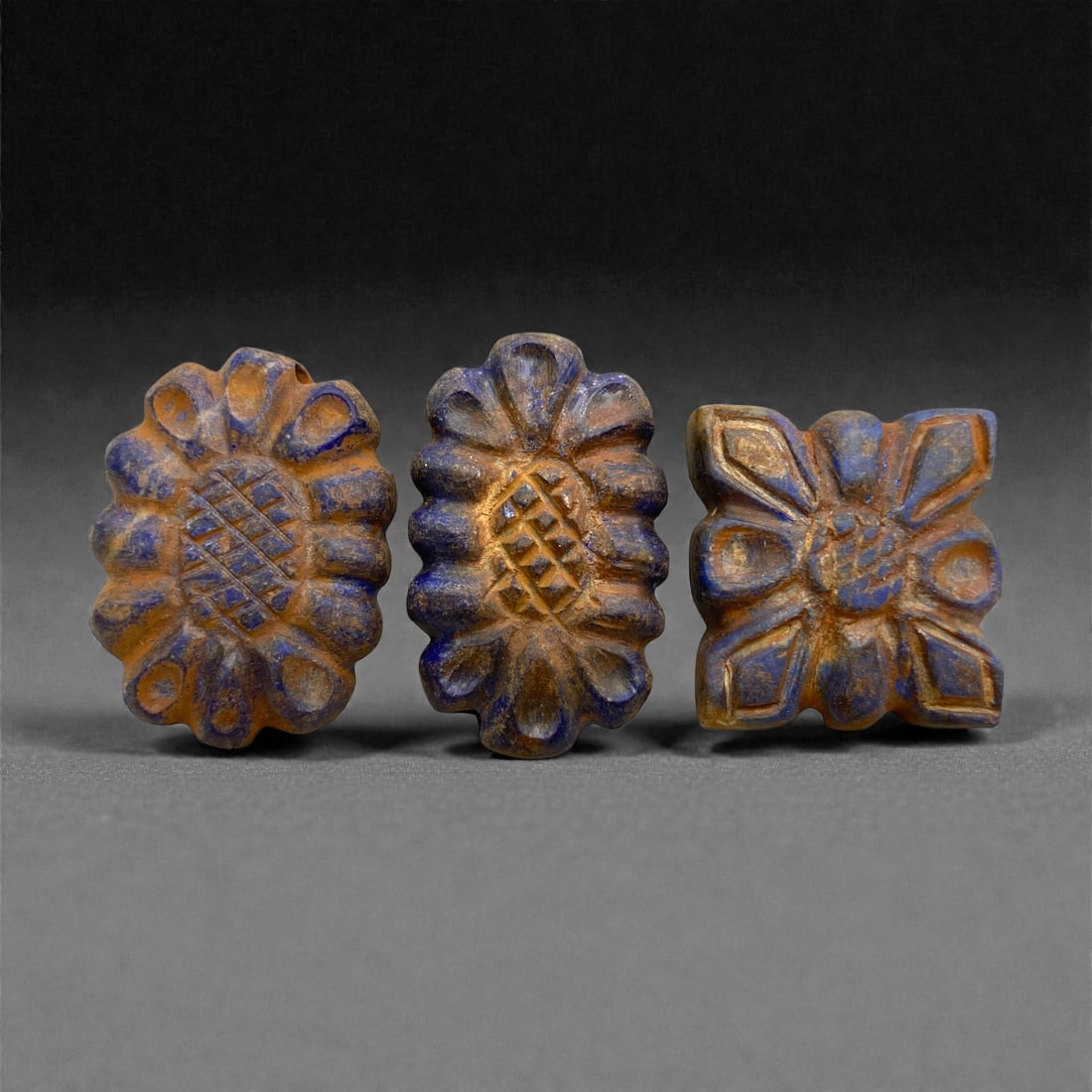 Achaemenid Lapis Lazuli Floral Beads, 5th-4th Century BCE, 100AA: Carved from blue lapis lazuli with surface weathering, the three beads comprise two oval rosettes with radiating rounded petals surrounding a central diamond-patterned field, and one square bead with 