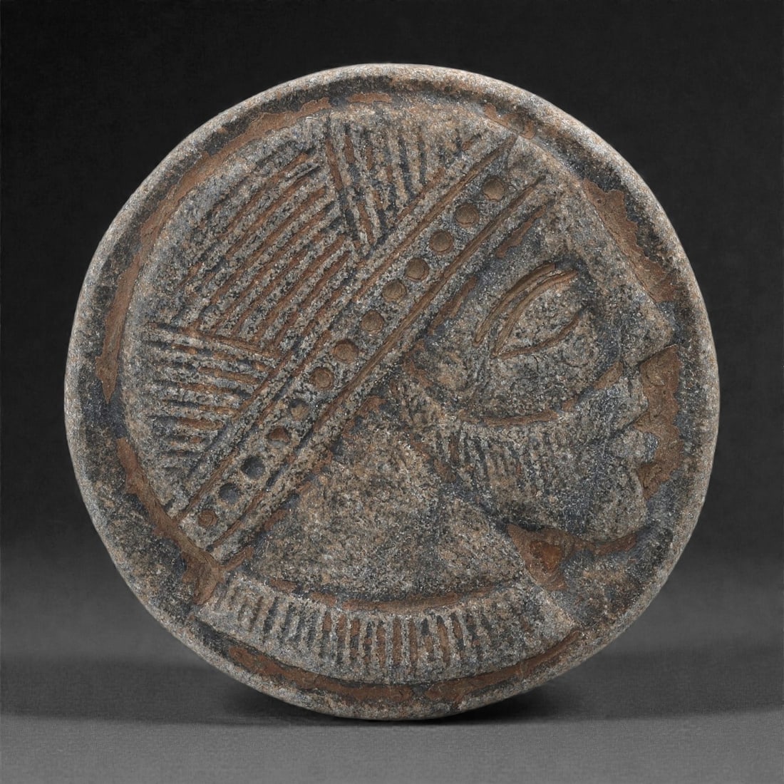 An Anatolian (Syro-Hittite) Steatite Plaque, Early 1st Millennium BCE, 78LR: Carved from grey-brown steatite, the circular plaque bears a profile head facing right, defined by a large eye with a recessed pupil, a pronounced nose, and an outlined jaw. The figure wears a broad h
