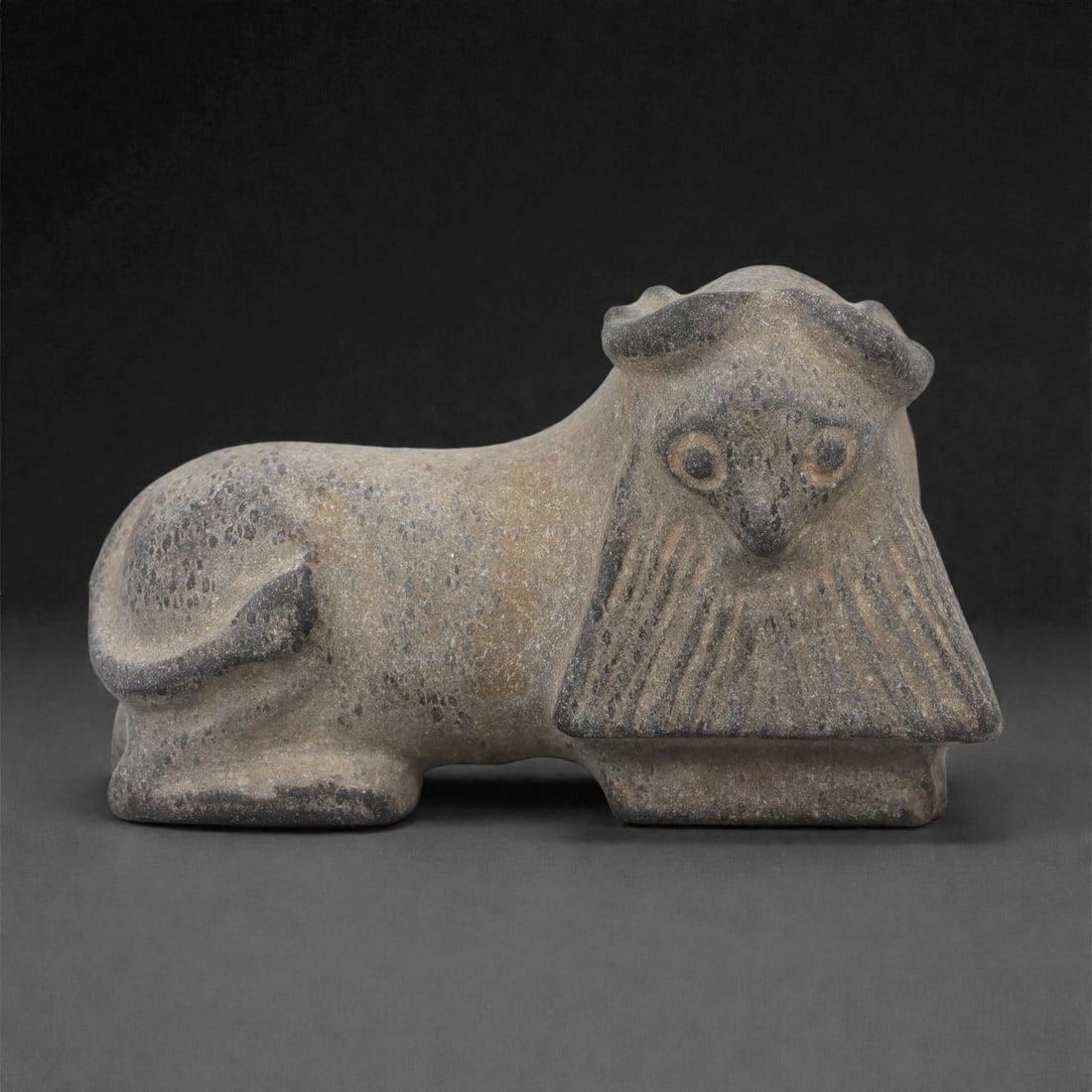 A Mesopotamian (Neo-Assyrian) Steatite Lamassu Figure, 9th-7th Century BCE, 16LR: Carved from grey steatite, the figure represents a recumbent lamassu with a compact body, curled tail, and short, block-like legs. The head is rendered frontally with circular recessed eyes, a short p