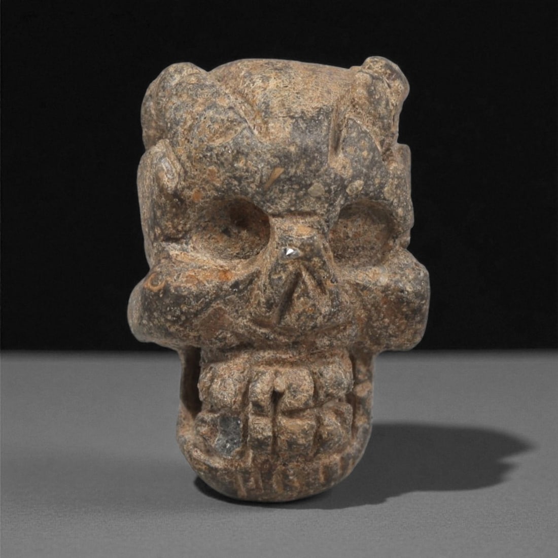 A Mediterranean Stone Skull Maskette, 12th-17th Century, 62LC: Carved from a mottled brown-grey stone with scattered lighter inclusions, the maskette is shaped in the form of a stylized skull. The eye sockets are deeply recessed, the nasal area is rendered as a t