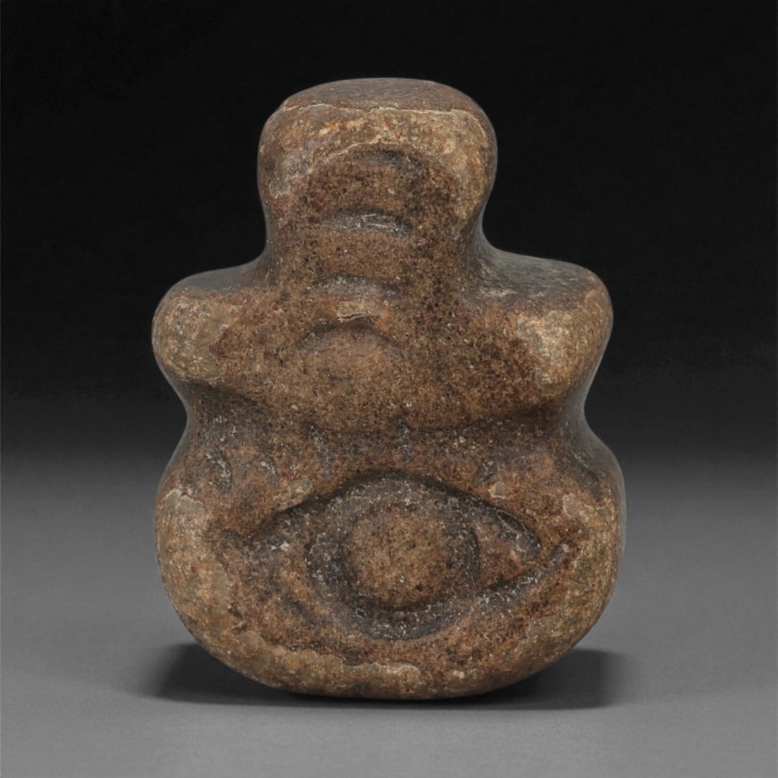 A Near Eastern Stone Eye-Motif Anthropomorphic Figurine, 3rd-2nd Millennium BCE, 73LB: Carved from a dark brown stone with lighter surface inclusions, the figurine is shaped with a compact lower body and a narrower upper section. The front is dominated by a large recessed circular eye e