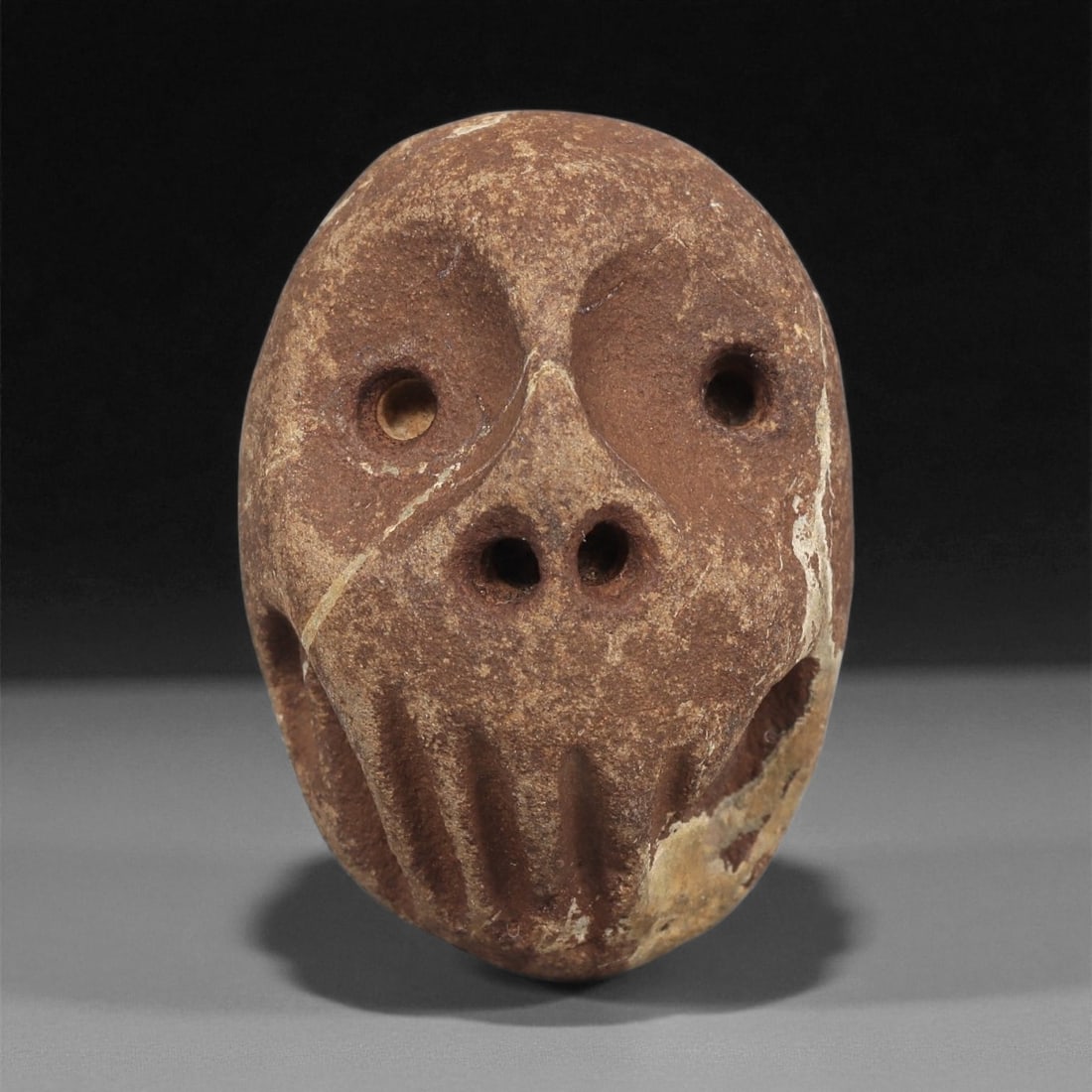 A Mediterranean Stone Maskette, 12th-17th Century, 23ZB: Carved from a reddish-brown stone with lighter surface veining, the maskette has an oval form tapering toward the chin. The facial features are executed in shallow relief, including circular recessed