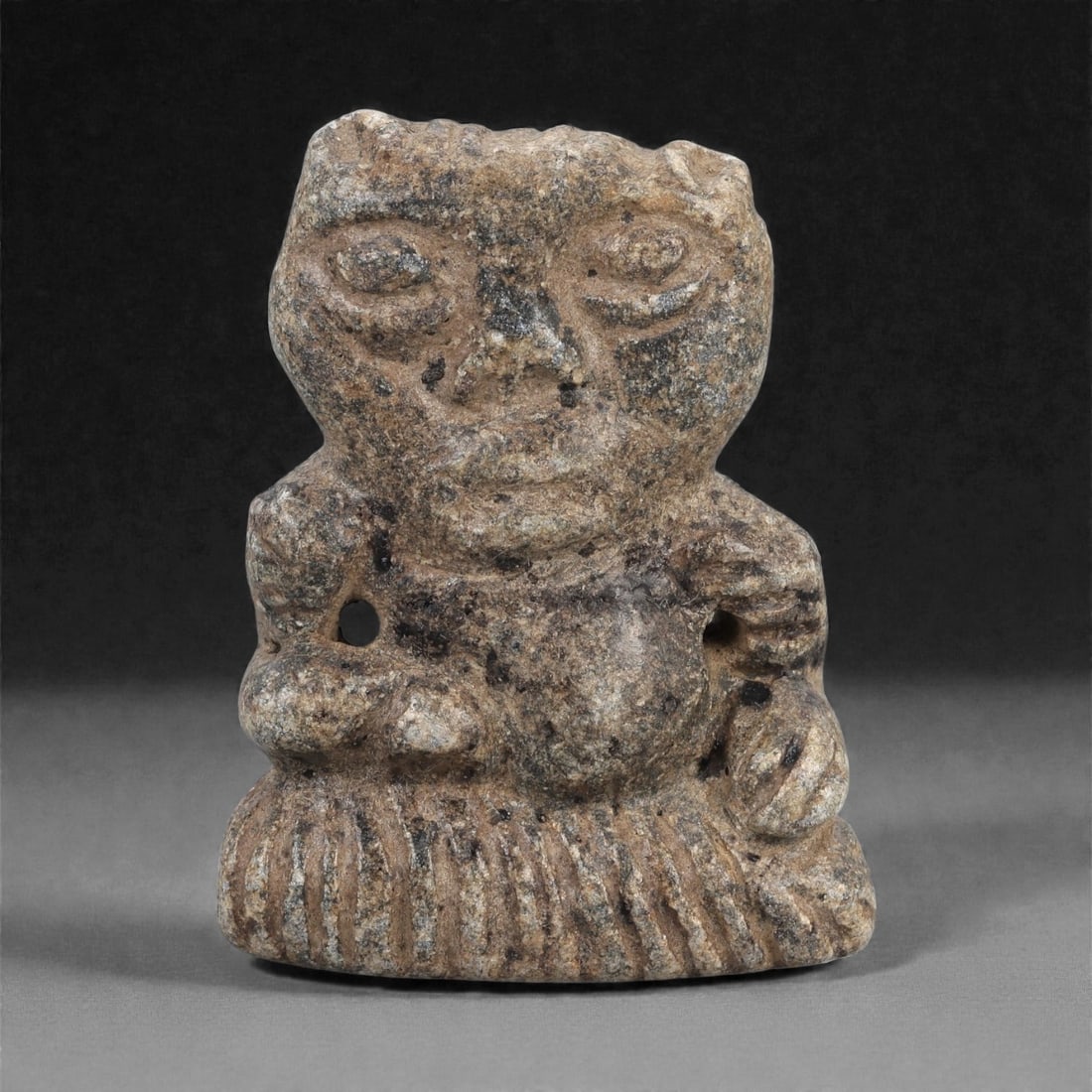 A Syro-Mesopotamian Schist Seated Figure, Early 2nd Millennium BCE, 98LJ: Carved from mottled grey-brown schist, the figure is shown seated on a flat, radiating base articulated with repeated vertical incisions. The body is compact, with bent arms positioned forward and han