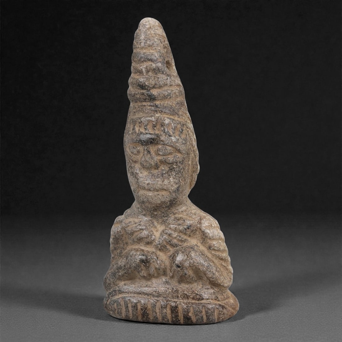A Syro-Hittite Schist Anthropomorphic Bust, Early 1st Millennium BCE, 94LJ: Carved from grey schist, the figure is rendered in a compact bust format with a tall conical headdress rising above a rounded face defined by incised eyes, a straight nose, and a horizontal mouth. The