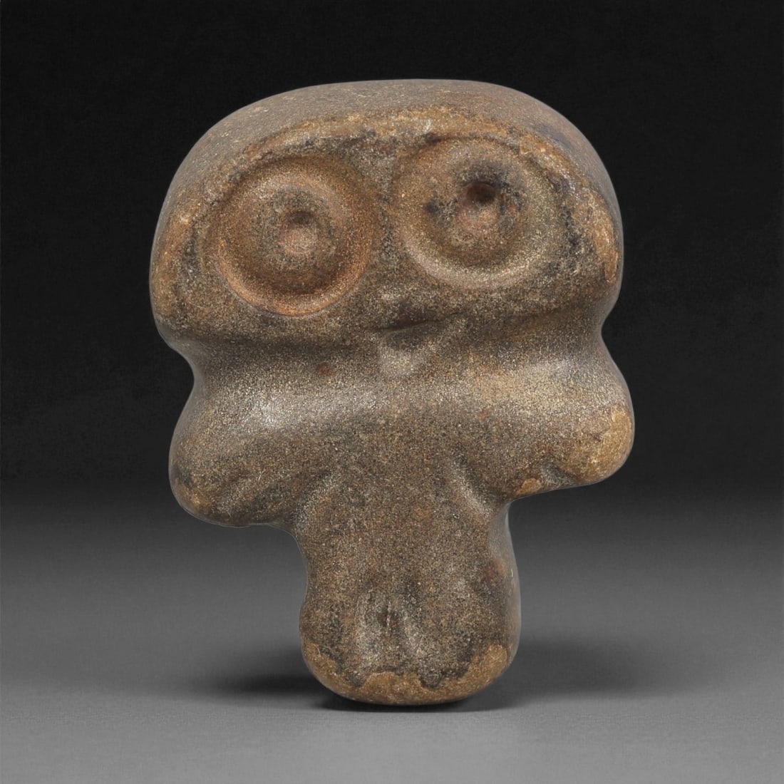 A Near Eastern Stone Anthropomorphic Figurine, 3rd-2nd Millennium BCE, 30ZB: Carved from a dark, fine-grained stone with light surface accretions, the figure presents a broad, rounded head dominated by large circular eyes formed through concentric recessed rings with central b