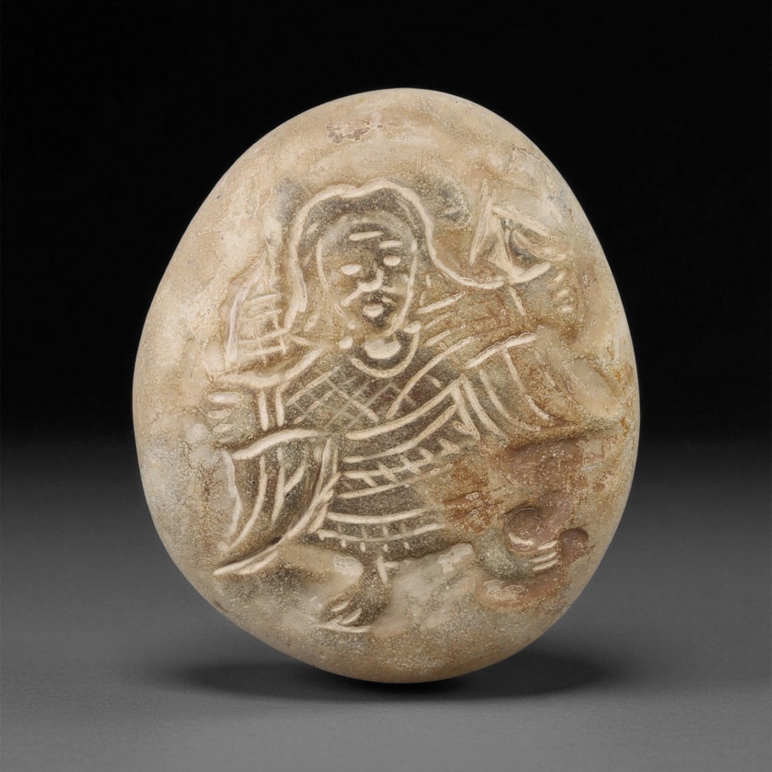 A Byzantine Stone Carved Pebble with Christian Figure, 6th-8th Century, 2LA: Carved from a light beige stone with natural surface inclusions, the pebble bears an incised depiction of a frontal human figure with delineated facial features, drapery indicated by cross-hatched and