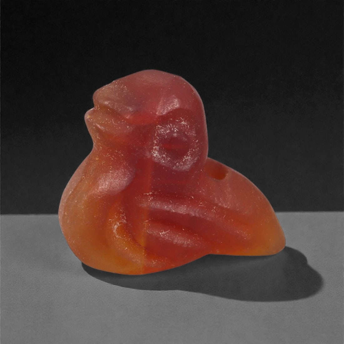 A Near Eastern Carnelian Bird Amulet, 1st Millennium BCE, 73LG: Carved from orange-red carnelian with natural internal color zoning, the amulet depicts a stylized bird with a compact body and rounded head. The wings are indicated through shallow linear carving alo