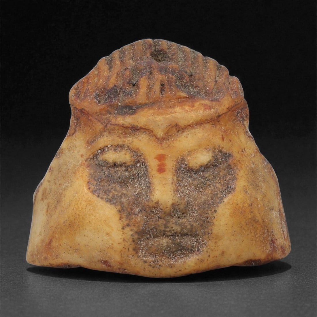 A Coptic Carved Bone Bust, 5th-7th Century CE, 6LJ: Carved from a light brown bone with darker surface accretions, the object is shaped as a small bust with a broad tapering base. The facial features are rendered in shallow relief, with the eyes, nose,