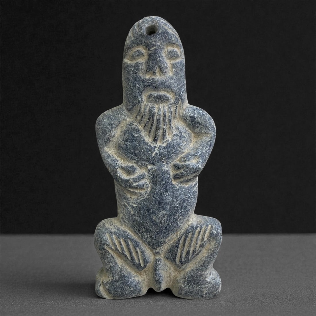 A Near Eastern Schist Anthropomorphic Figurine, 3rd-2nd Millennium BCE, 36g, 21EE: Carved from dark grey schist with lighter inclusions, the figurine is modeled in a crouching posture with the arms brought forward across the torso. The face is rendered with incised eyes, nose, and b