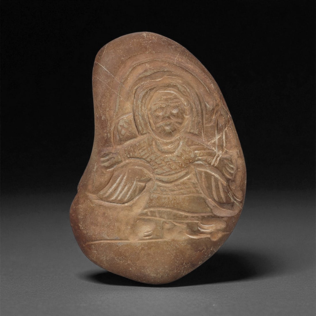 A Byzantine Stone Carved Pebble with Christian Figure, 6th-8th Century CE, 88LD: Carved from a naturally rounded brown pebble with fine surface patination, the object bears a shallow relief of a frontal haloed figure. The figure is depicted with outstretched arms, the drapery rend