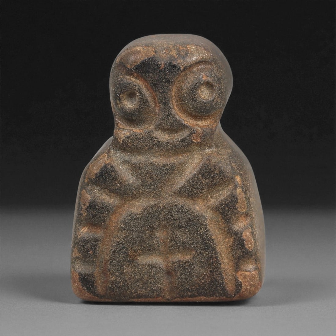 A Near Eastern Stone Anthropomorphic Figurine, 4th-3rd Millennium BCE, 94LD: Carved from a dark brown stone with lighter surface abrasions, the figurine is modeled with a rounded head, large recessed circular eyes, and a small incised mouth. The upper body is delineated by inc
