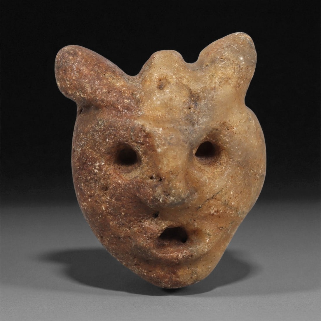 A Mediterranean Stone Maskette, 12th-17th Century, 2LC: Carved from a light brown stone with scattered darker inclusions, the maskette presents a rounded facial form tapering to a narrow chin. Two prominent upper projections, possibly stylized ears or horn