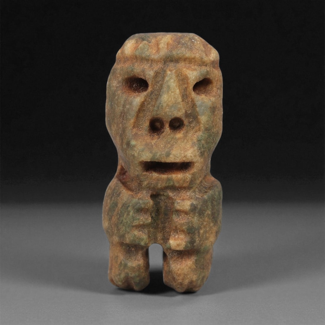 A Taino Carved Stone Idol, Late Ceramic Age, circa 1200-1500 CE, 73LD: Carved from mottled brown-green stone with areas of surface weathering, the figure is shown standing with short bifurcated legs and a compact body. The head is rounded with recessed circular eyes, dri