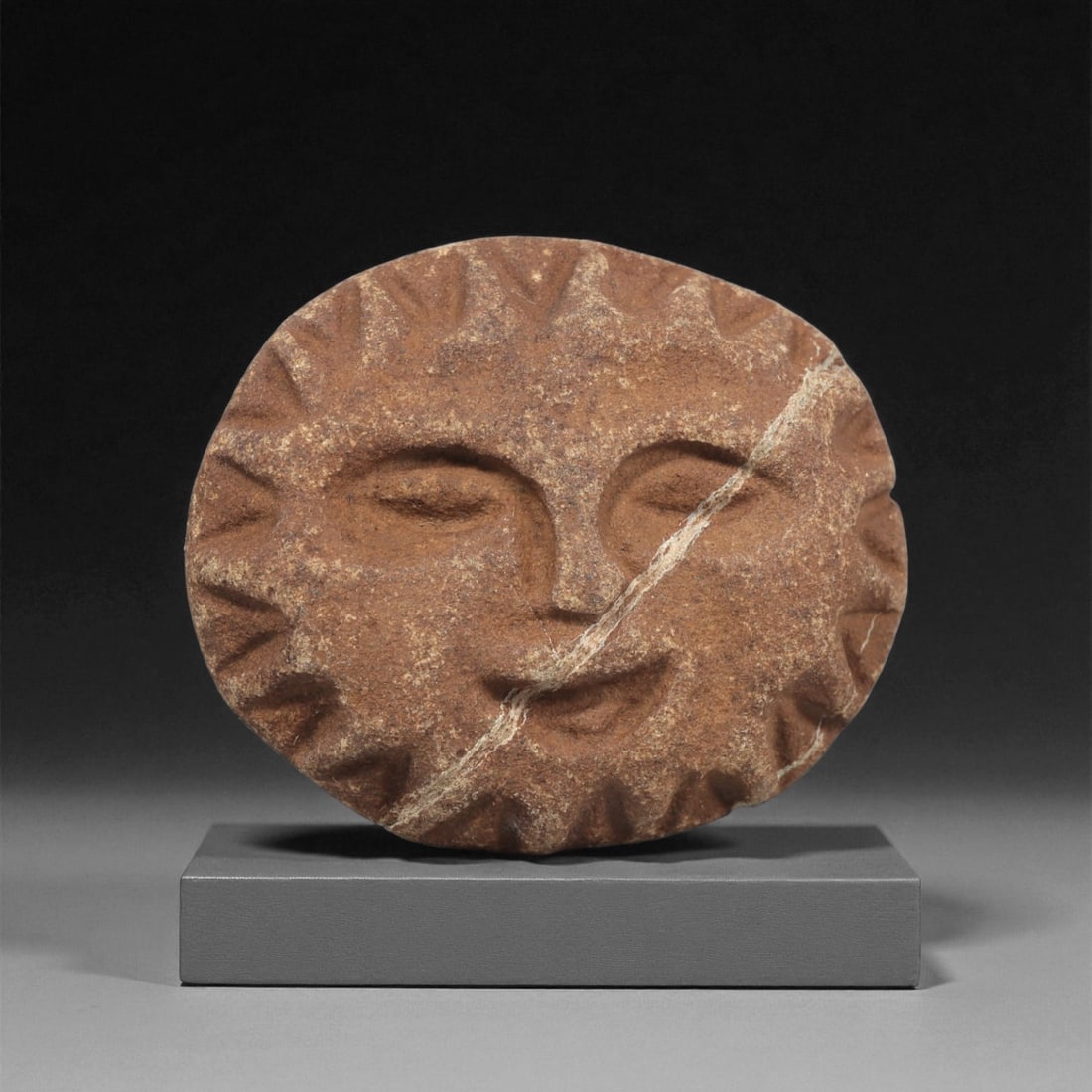 A Mediterranean Stone Sun-Face Maskette, 12th-17th Century, 83LC: Carved from a reddish-brown stone with lighter mineral veining, the maskette is shaped as a circular face encircled by short triangular rays cut in shallow relief. The interior features recessed eyes