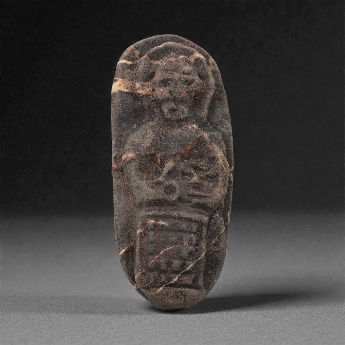 A Byzantine Stone Carved Figural Pebble, 6th-8th Century CE, 59LH: Carved from a dark brown pebble with lighter veining, the object is naturally elongated and rounded at the upper end. The front bears a shallow relief depicting a frontal figure with a circular head f