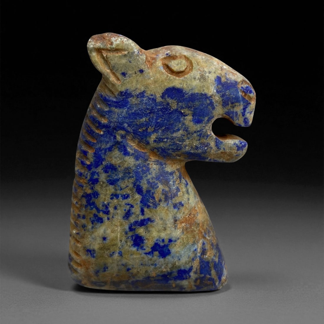 A Near Eastern Lapis Lazuli Feline Bust Effigy, 2nd Millennium BCE, 90g, 29EE: Carved from lapis lazuli with mottled blue and tan surface tones, the effigy is shaped as a feline head and upper torso with the mouth open and the snout projecting forward. Incised lines define the e