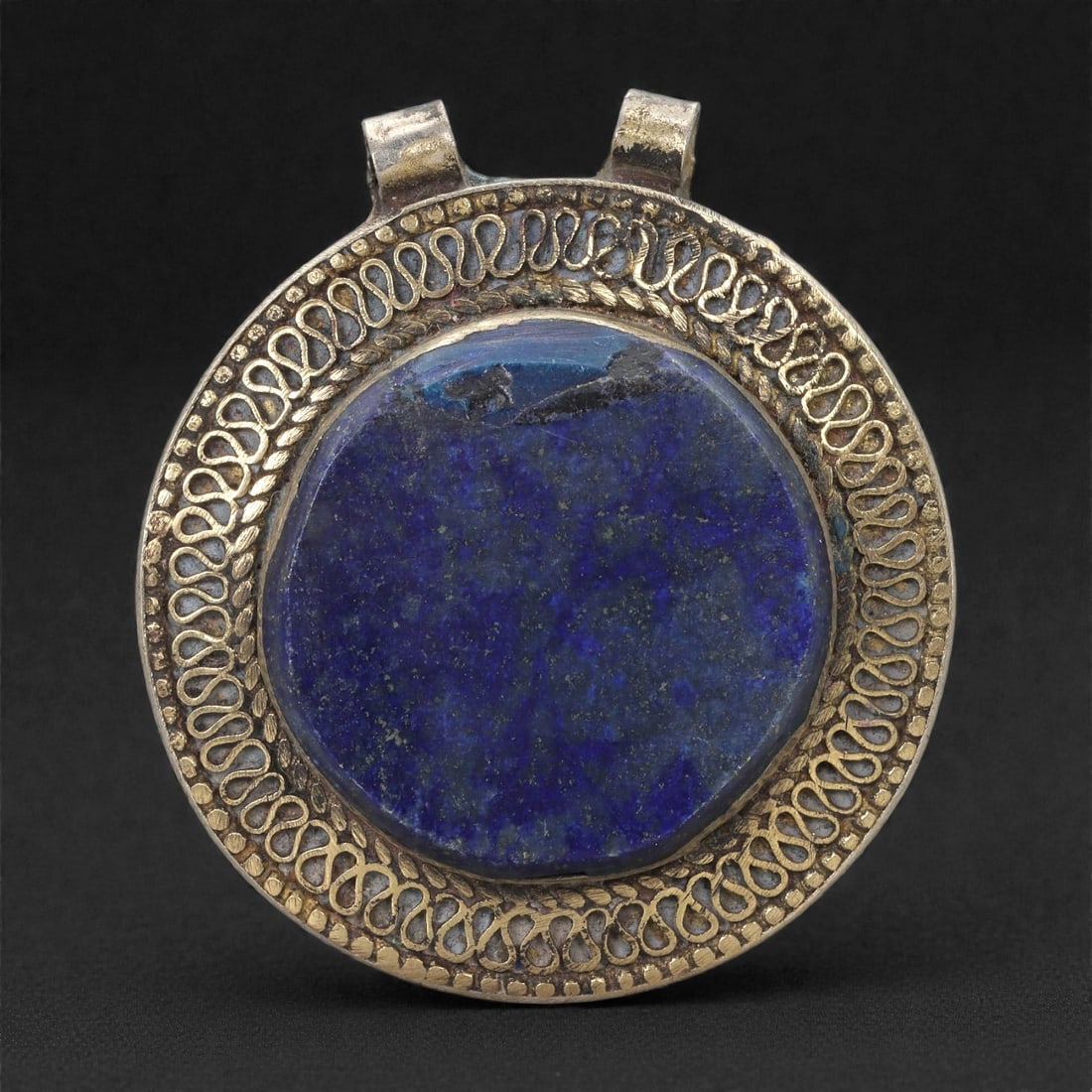 Himalayan Style Brass Pendant - Lapis Cabochon, 1XF: This circular brass pendant is dominated by a large lapis cabochon whose surface shows natural mottling and areas of wear, giving the stone a subdued, matte character. The cabochon is enclosed within