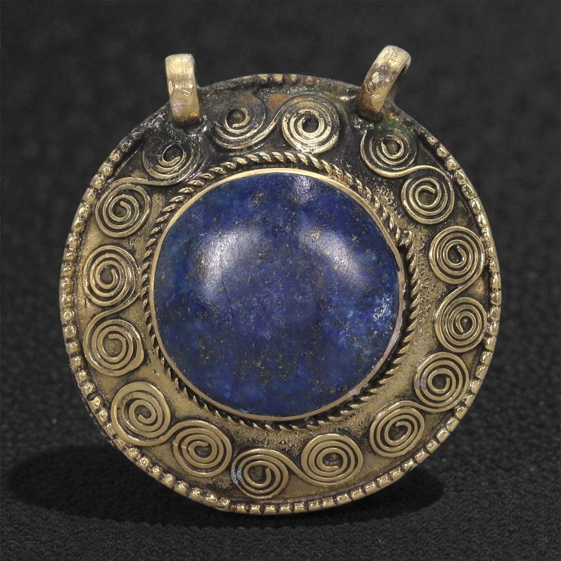 Himalayan Brass Pendant - Deep Blue Lapis-Spiral Motif, 44XF: This round brass pendant centers on a deep blue cabochon whose surface shows natural mottling and subtle variations in tone. The stone is framed by a twisted-wire bezel, followed by a ring of meticulo