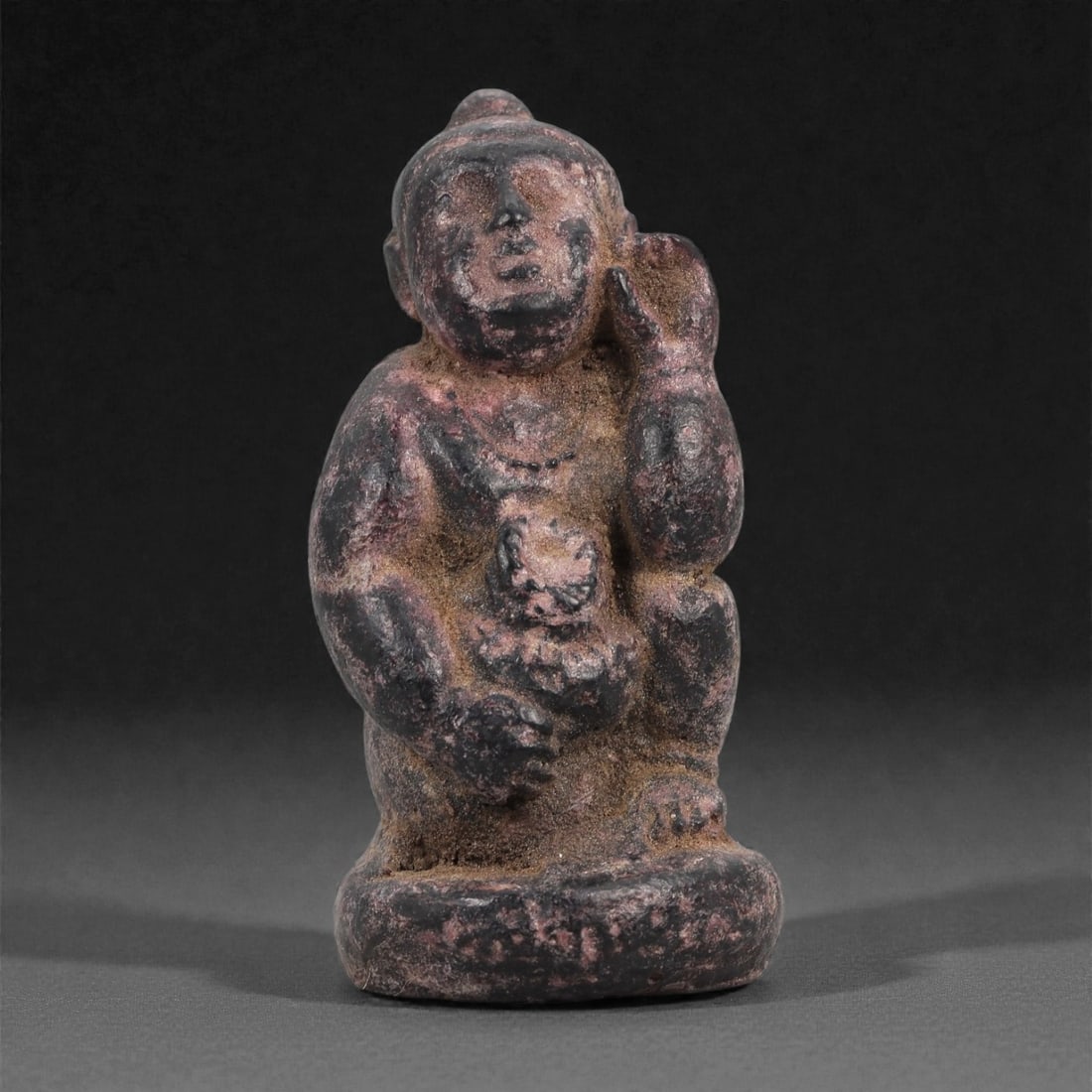 Hard Resin Seated Child Figure - South Asian Iconographic Figurine, 32D: Formed in hard resin with a dark surface bearing reddish pigmentation, this small figurine depicts a seated child-like figure resting one hand against the side of the head while the other holds a roun