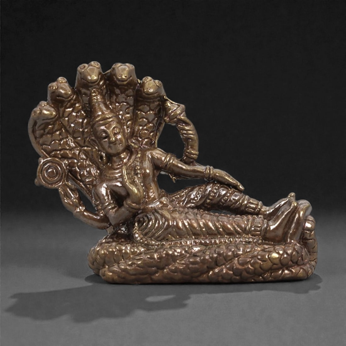 Bronze Reclining Deity on Multi-Headed Serpent - Hindu Naga Icon Figurine, 21D: Cast in bronze with a warm reflective surface, this compact sculpture portrays a reclining deity resting upon the coiled body of a multi-headed serpent. The figure is shown in a relaxed pose, one arm