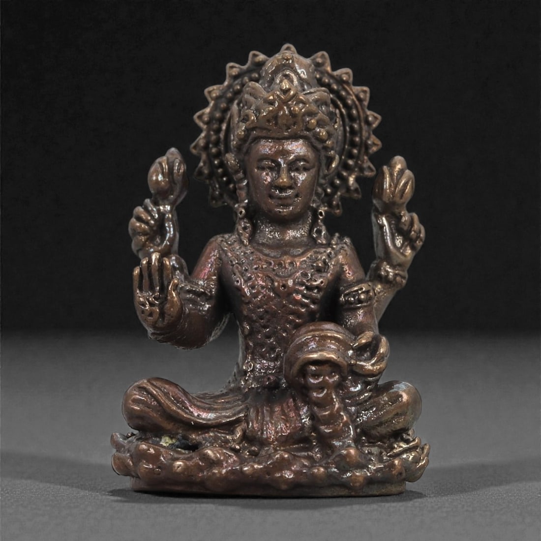 Bronze Four-Armed Deity Idol - Hindu Iconographic Figurine, 18D: Cast in bronze with a warm, subtly reflective surface, this miniature sculpture depicts a four-armed deity seated cross-legged upon a lotus-form base. The figure wears an ornate headdress that radiate