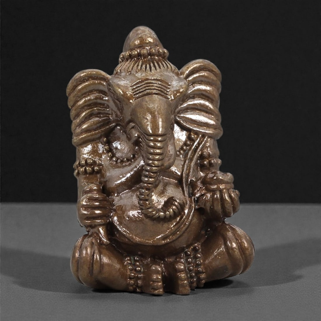 Bronze Seated Ganesha Idol - Hindu Deity Figurine, 14D: Cast in bronze with a warm, polished surface, this compact figurine depicts Ganesha seated cross-legged in a composed frontal posture. The elephant head is characterized by a long curved trunk, flarin