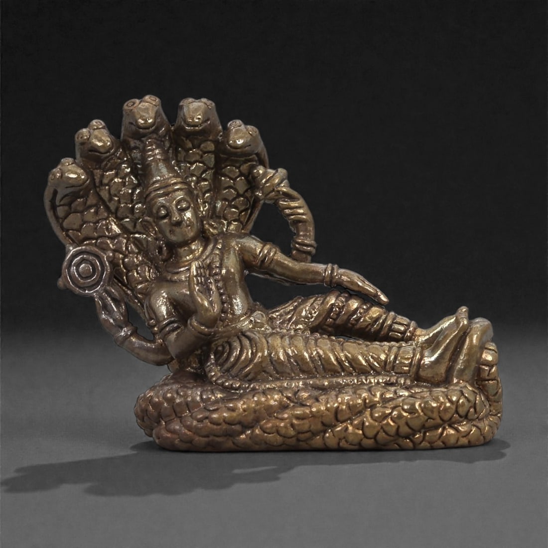 Bronze Reclining Deity on Multi-Headed Serpent - Hindu Naga Icon Figurine, 1D: Cast in bronze with a warm reflective surface, this miniature sculpture depicts a reclining deity supported by the coiled body of a multi-headed serpent. The figure rests on one arm while the other ho