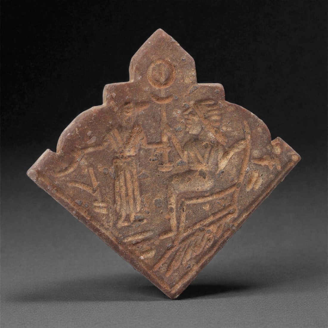 A Sasanian Stone Plaque Amulet, 3rd-7th Century CE, 63LR: Carved from brown stone, the plaque is shaped with a fan-like lower body and a stepped, pointed upper edge. The front surface bears a shallow relief scene depicting a seated figure facing a standing a