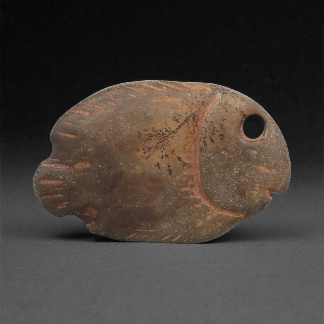 A Near Eastern Stone Fish Figurine, 3rd Millennium BCE, 29LF: Carved from a brown stone with darker surface mottling and natural inclusions, the object is shaped as a stylized fish with a rounded body, a notched tail, and an incised mouth. A drilled circular eye