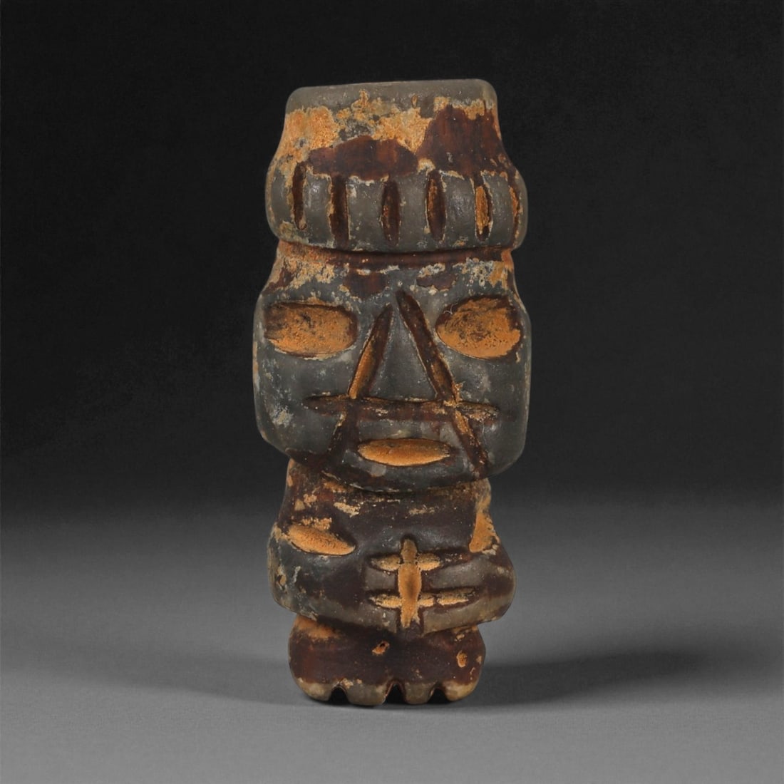 A Taino Carved Stone Idol, Late Pre-Columbian Period, Circa 1000-1500 CE, 21ZB: Carved from dark brown stone with lighter surface encrustations, the figure stands upright with short legs and a compact torso. The head is surmounted by a band incised with vertical grooves, while th