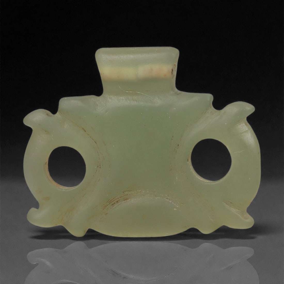 An Islamic Jade Openwork Amulet, Medieval Period, 12th-14th Century CE, 94LP: Carved from pale green translucent jade, the amulet is shaped as a compact rectangular body flanked by two rounded lateral extensions, each pierced with a large circular aperture. Additional small pro