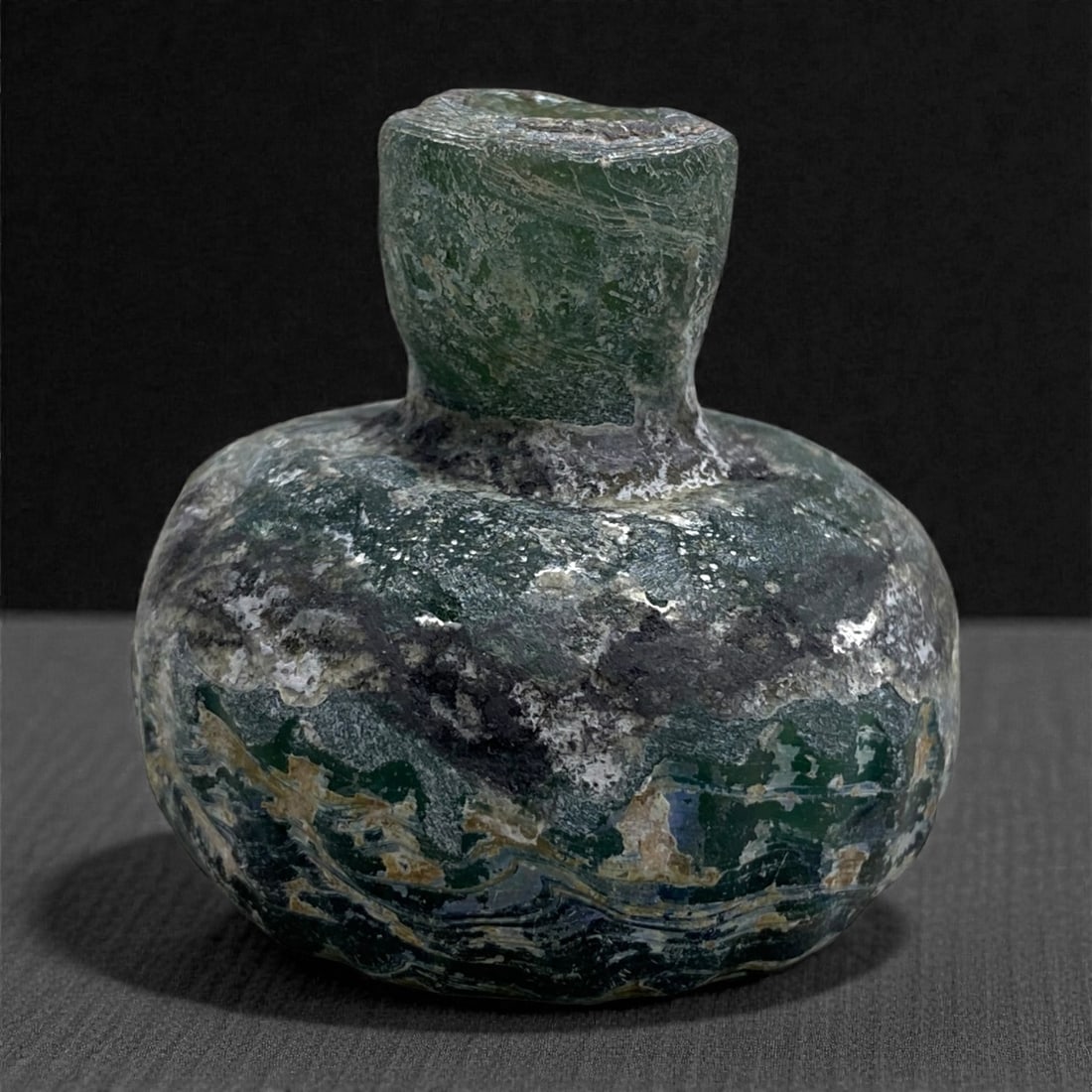 A Roman Levantine Glass Aryballos, 1st Century CE, 79 g, 29AA: Blown from dark green glass with heavy weathering and mineral accretion, the vessel has a globular body rising to a short cylindrical neck with a slightly flattened rim. The form corresponds to small 