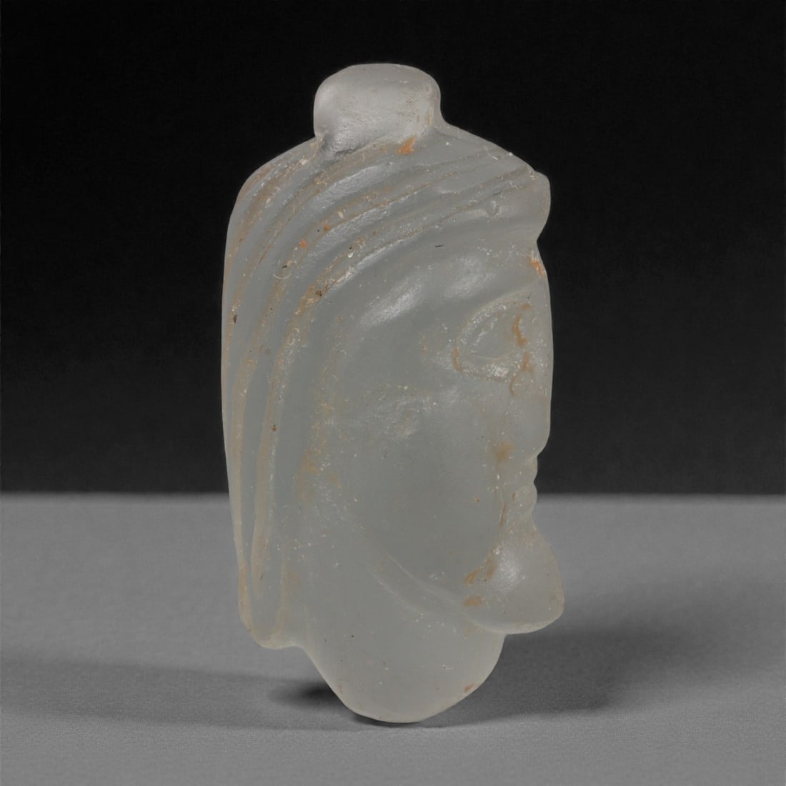 A Sasanian Glass Noble-Head Amulet, 3rd-7th Century CE, 37LQ: Carved from translucent pale glass, the amulet depicts a male head in profile with a pronounced brow, almond-shaped eye, and a slightly projecting chin. The hair is rendered by a series of parallel in