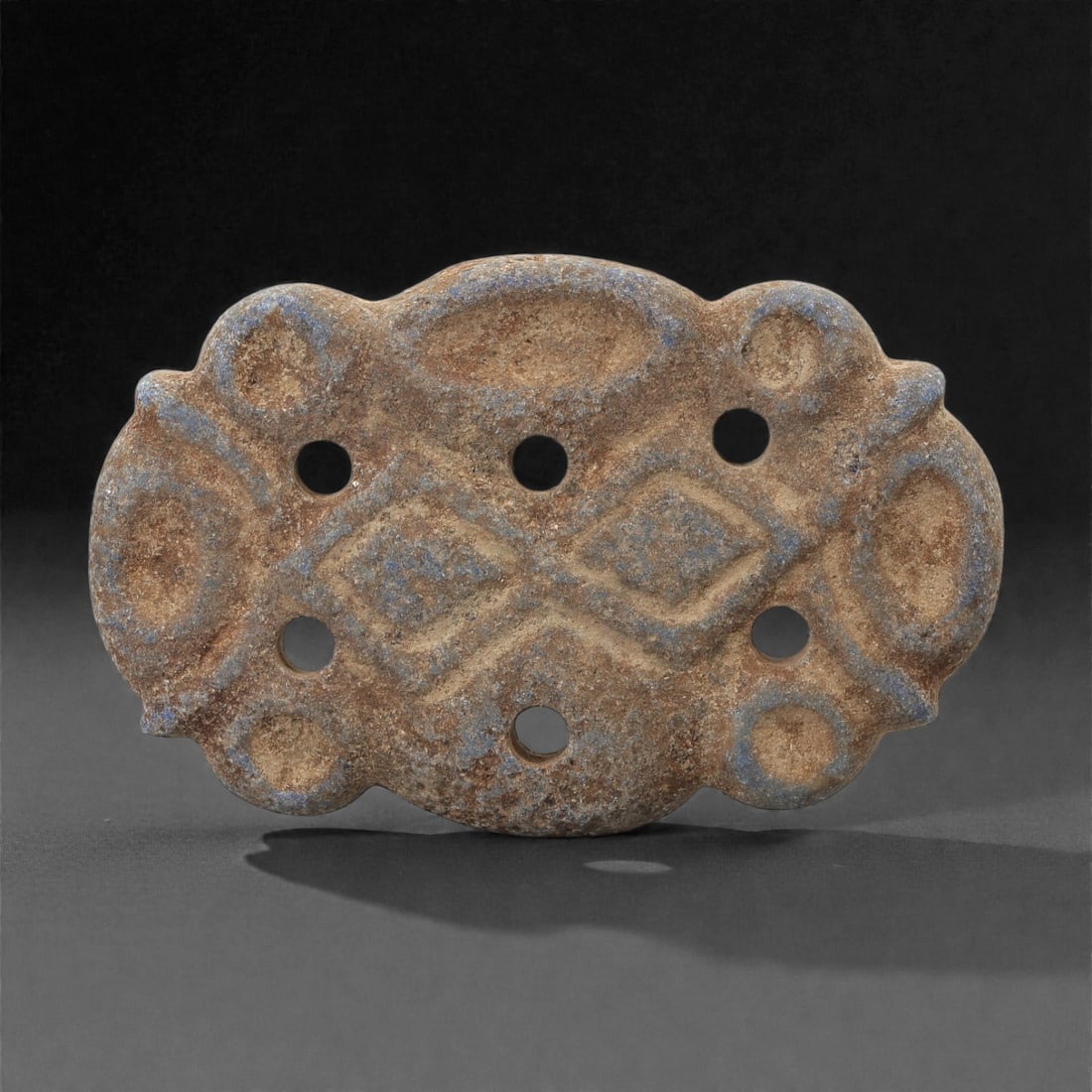 A Parthian Lapis Lazuli Openwork Amulet, 1st Century BCE-2nd Century CE, 30LR: Carved from lapis lazuli with brown archaeological surface accretions, the amulet is fashioned as a flat plaque with scalloped edges and a central field composed of raised lozenge and oval motifs arra