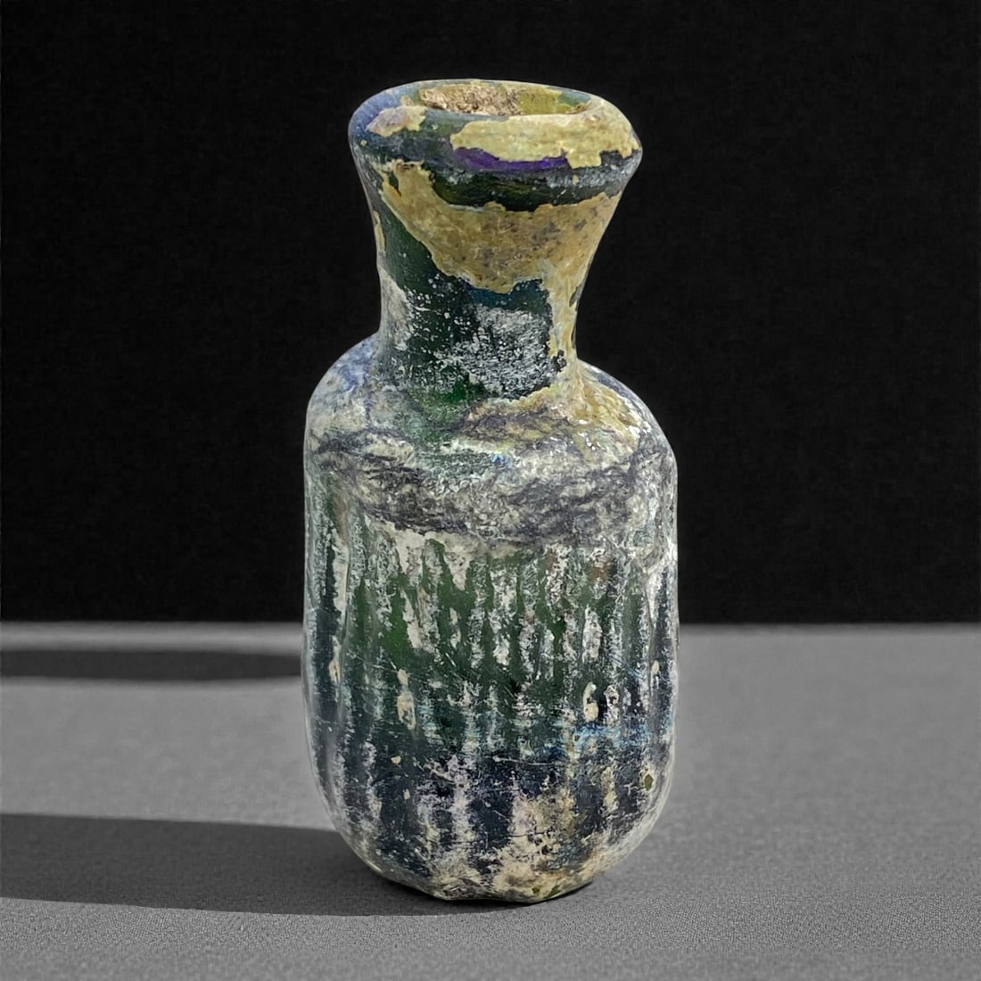 A Roman Levantine Mold-Blown Glass Unguentarium, 1st-2nd Century CE, 49 g, 53AA: Blown from dark green glass now obscured by iridescence, pitting, and heavy burial accretion, the vessel is formed with a rounded body transitioning into a short, slightly tapering neck with a thicken