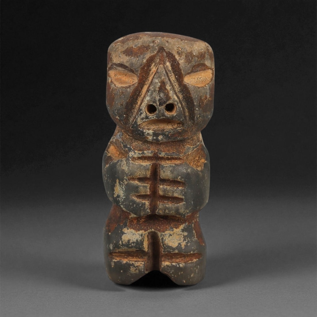 A Taino Carved Stone Idol, Late Pre-Columbian Period, Circa 1000-1500 CE, 37ZA: Carved from mottled dark brown and grey stone with lighter surface abrasions, the figure stands upright with short legs and a compact torso. The head is modeled with recessed oval eyes, a sharply tria
