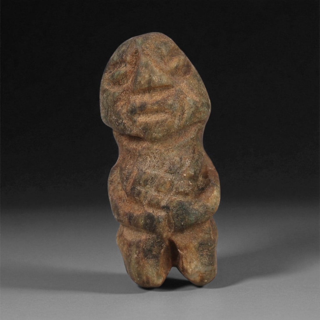 A Taino Carved Stone Idol, Late Pre-Columbian Period, Circa 1000-1500 CE, 87LF: Carved from a mottled brown-green stone with light surface weathering, the figure stands with the legs divided by a narrow central groove. The torso is modeled with a series of incised chevron-like li