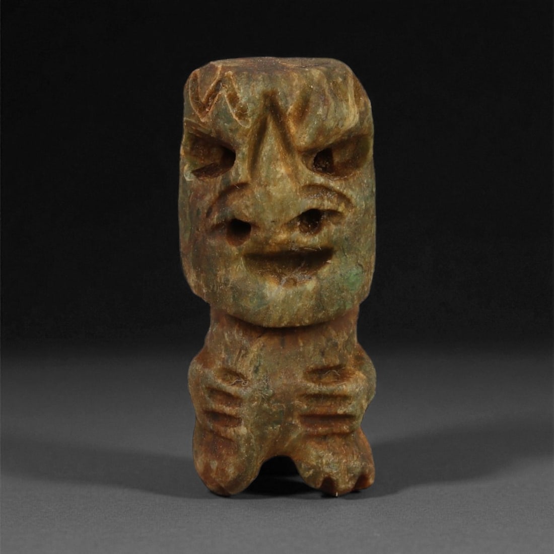 A Taino Carved Stone Idol, Late Pre-Columbian Period, Circa 1000-1500 CE, 56LA: Carved from a mottled brown-green stone with surface accretions, the figure is modeled with a large squared head and compact body. The face features recessed eyes beneath sharply incised brows, a prom
