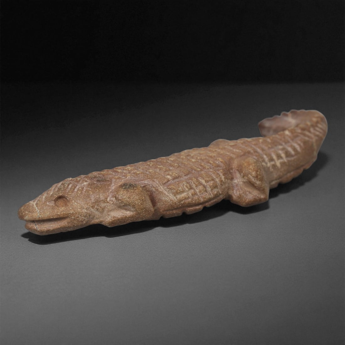 An Egyptian Stone Crocodile Figure, Late Period, 664-332 BCE, 3LA: Carved from brown stone with lighter granular inclusions, the figure depicts a crocodile in elongated form with a tapering tail. The head is rendered with a gently rounded snout, incised mouth line, a