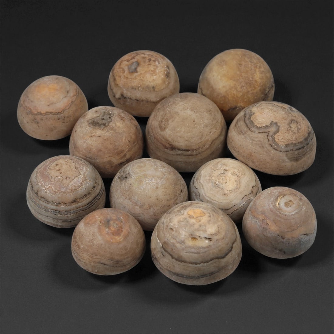 Tibetan Stone Luk Mik Beads, Possibly 19th-20th Century, 56LE: Worked from banded stone with natural concentric veining, the group comprises twelve hemispherical bead forms, each shaped with a rounded dome and a flattened base. The surfaces show layered mineral a