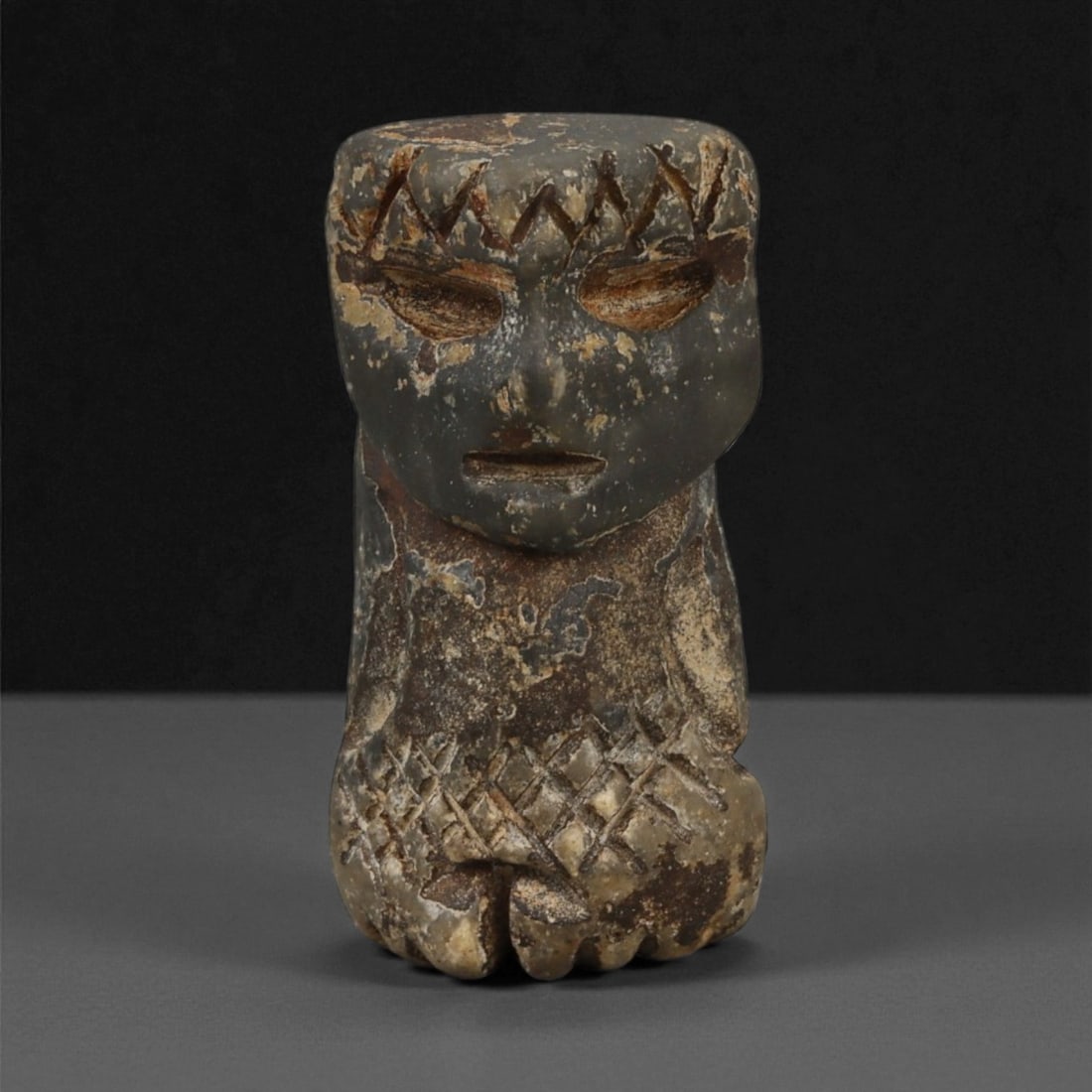 A Taino Carved Stone Idol, Late Pre-Columbian Period, Circa 1000-1500 CE, 61LA: Carved from a dark stone with lighter surface encrustations, the figure is modeled with a broad, squared head and compact lower body. The face bears recessed oval eyes, a straight incised line for the