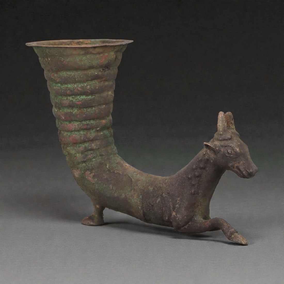 An Achaemenid Bronze Rhyton with Recumbent Ibex, 5th-4th Century BCE, 72LP: Cast in bronze as a small rhyton, the vessel is formed with a flaring, ribbed spout rising from the back of a recumbent ibex. The animal is rendered with an elongated neck, short projecting muzzle, an