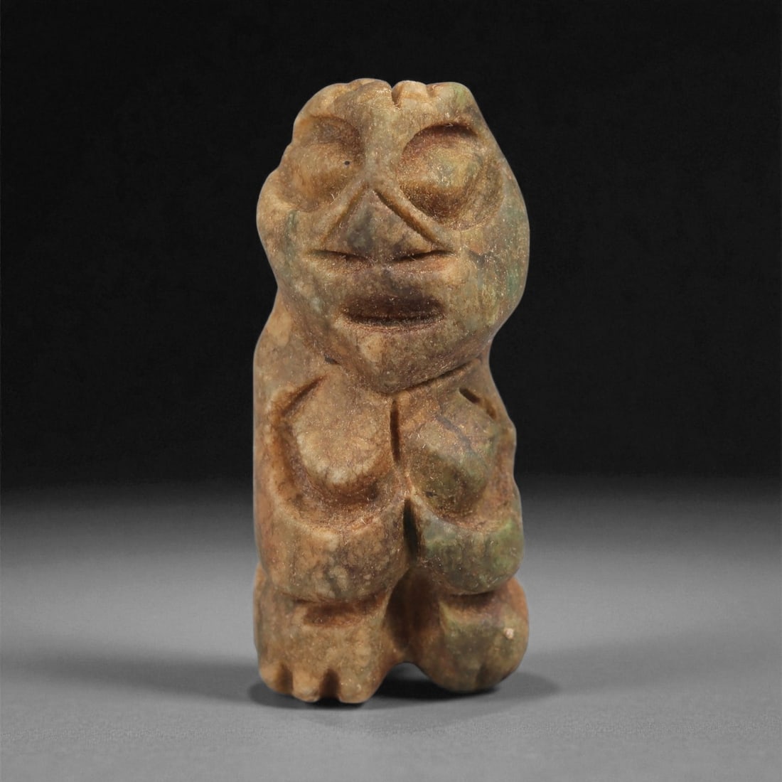 A Taino Carved Stone Idol, Late Ceramic Age, circa 1200-1500 CE, 48LD: Carved from mottled brown-green stone with light surface weathering, the figure is shown standing with short legs and a compact body. The head is rounded with recessed eyes, an incised triangular nose