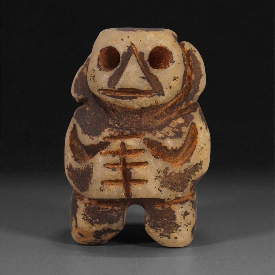 A Taino Carved Stone Idol, Late Pre-Columbian Period, Circa 1000-1500 CE, 47ZA: Carved from mottled cream and brown stone, the figure stands with short bifurcated legs and a broad torso. The head is modeled with two large drilled circular eyes, a triangular nose rendered in low r