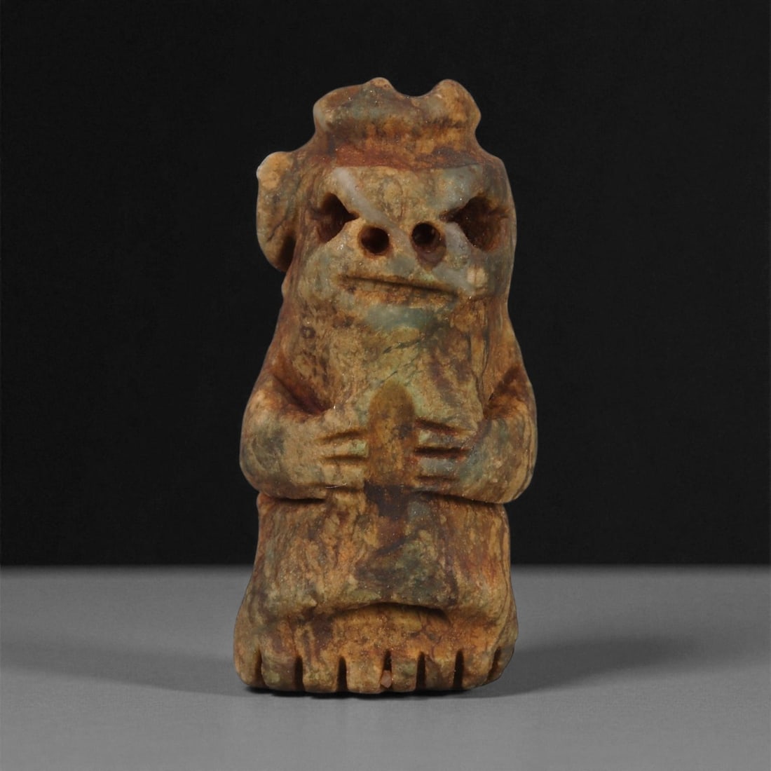 A Taino Carved Stone Idol, Late Pre-Columbian Period, Circa 1000-1500 CE, 57ZA: Carved from mottled brown and green stone, the figure stands upright with short legs and arms held at the abdomen. The head is formed with a pronounced brow, drilled circular eyes, and a straight inci