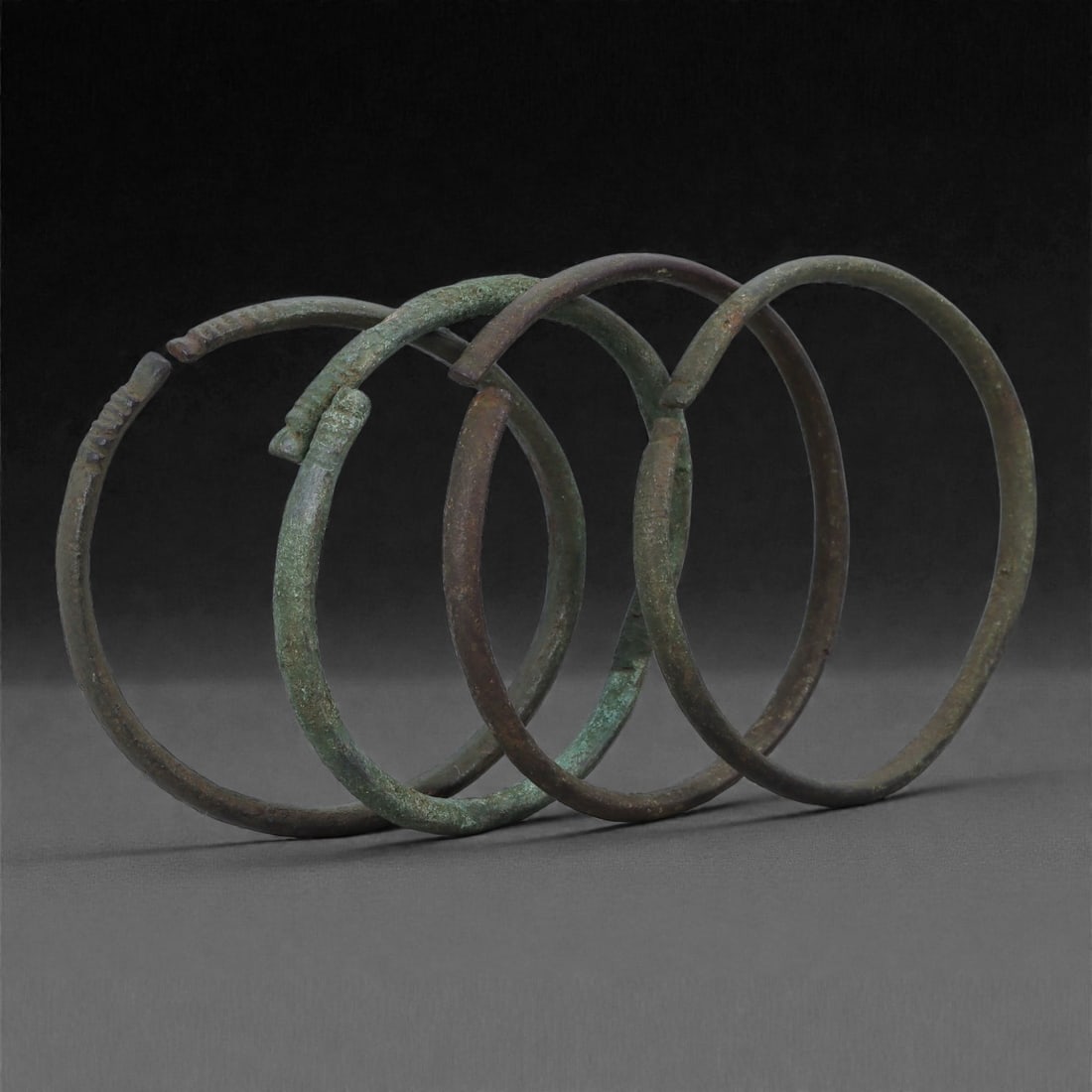 A Group of Roman Bronze Bracelets, 1st-3rd Century CE, 54H: Cast in bronze with slender circular-section profiles, the group comprises four open bracelets displaying surface patination in green and brown tones. Each is formed from a thin rod bent into an annul