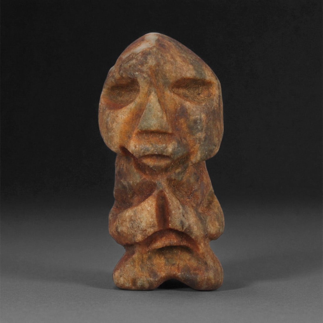A Taino Carved Stone Idol, Late Pre-Columbian Period, Circa 1000-1500 CE, 87ZA: Carved from mottled brown and grey stone with scattered surface accretions, the figure stands upright with short legs and a compact torso. The head is formed with recessed oval eyes, a sharply triangu